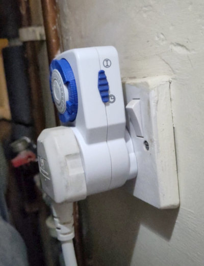 A UK electrical plug plugged into a wall socket via a mechnical timer. Override switch is shown on the side.